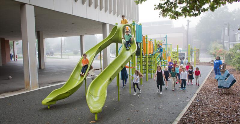 School kids playing in the misting rain on a playground with two limon colored slides in the foreground.