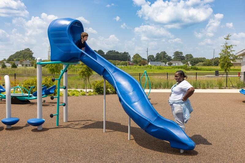 A boy prepares to slide down a standalone Rushwinder slide on a playground while a woman watches from below