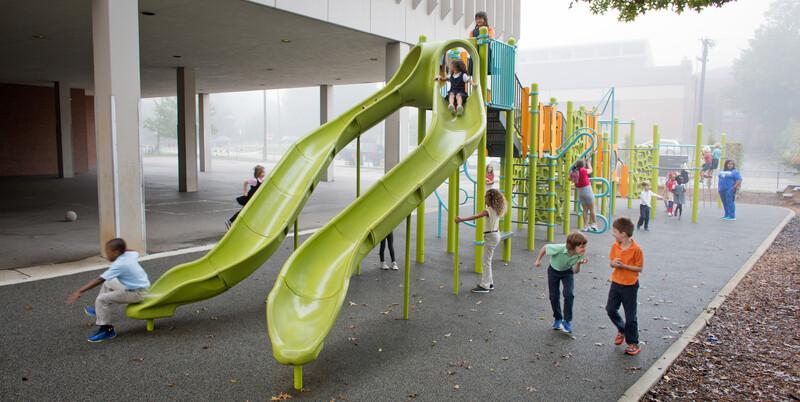 School kids playing in the misting rain on a playground with two limon colored slides in the foreground. 
