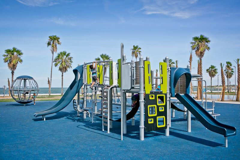 Boy climbing on second level of Smart Play structure in McCaughan Park. The park sits by the beach. Palm trees stretch up to the big blue sky.
