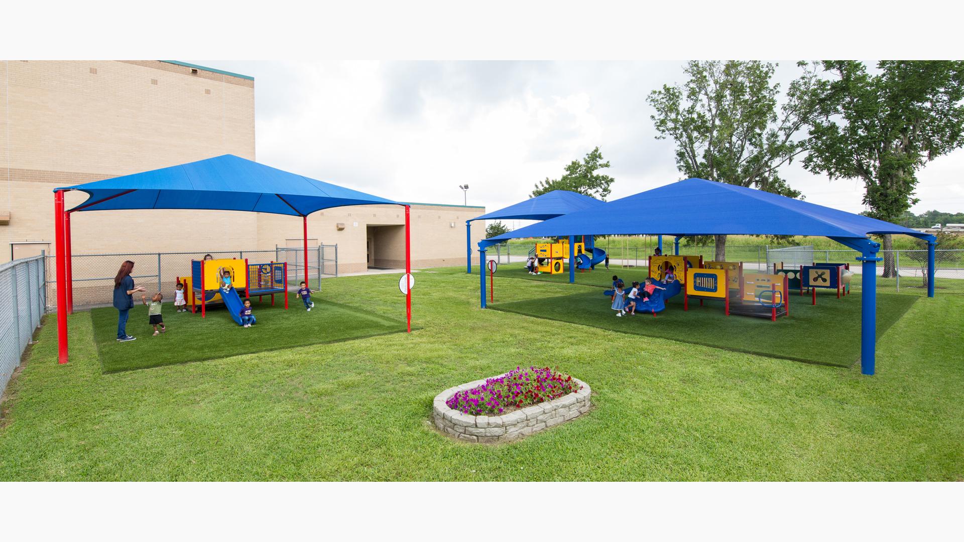 Childcare-aged kids playing on primary-colored playground equipment. Three separate playground areas covered by blue shade.