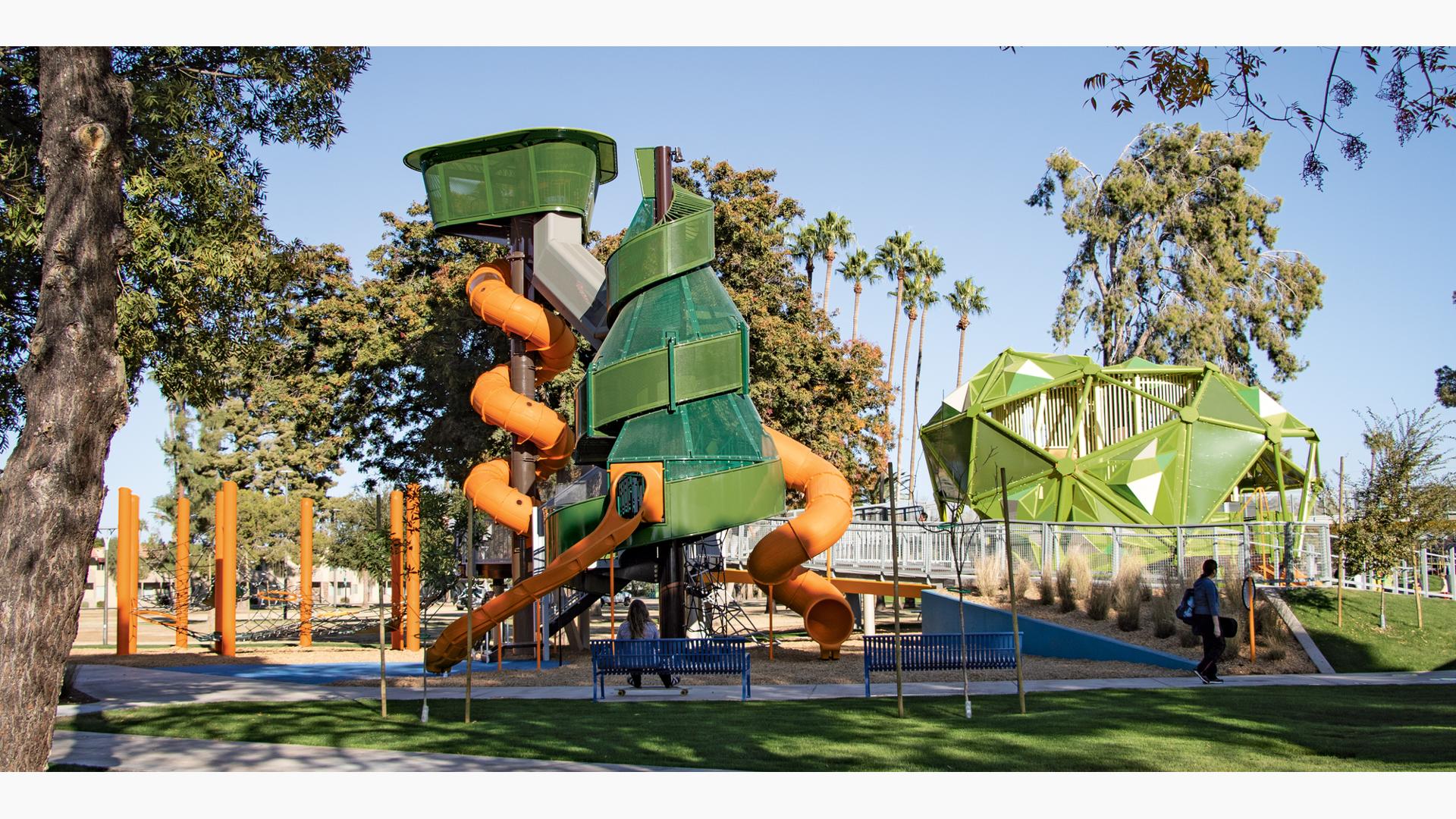 Woman sitting on bench in front of play structure as man person with skateboard walks by on a sunny day.