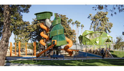 Woman sitting on bench in front of play structure as man person with skateboard walks by on a sunny day.