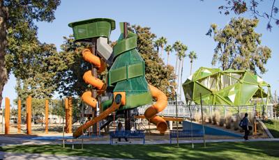 Woman sitting on bench in front of play structure as man person with skateboard walks by on a sunny day.