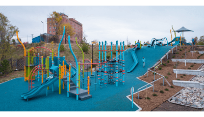  A large playground sits at the bottom of a small hill side leading up to a secondary play structure. A large blue slide cascades down the hillside connecting the two play areas.