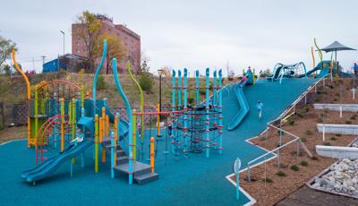  A large playground sits at the bottom of a small hill side leading up to a secondary play structure. A large blue slide cascades down the hillside connecting the two play areas.