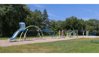 Children play on playground activities strung in a line for a continues play experience with a lush tree line in the background. 