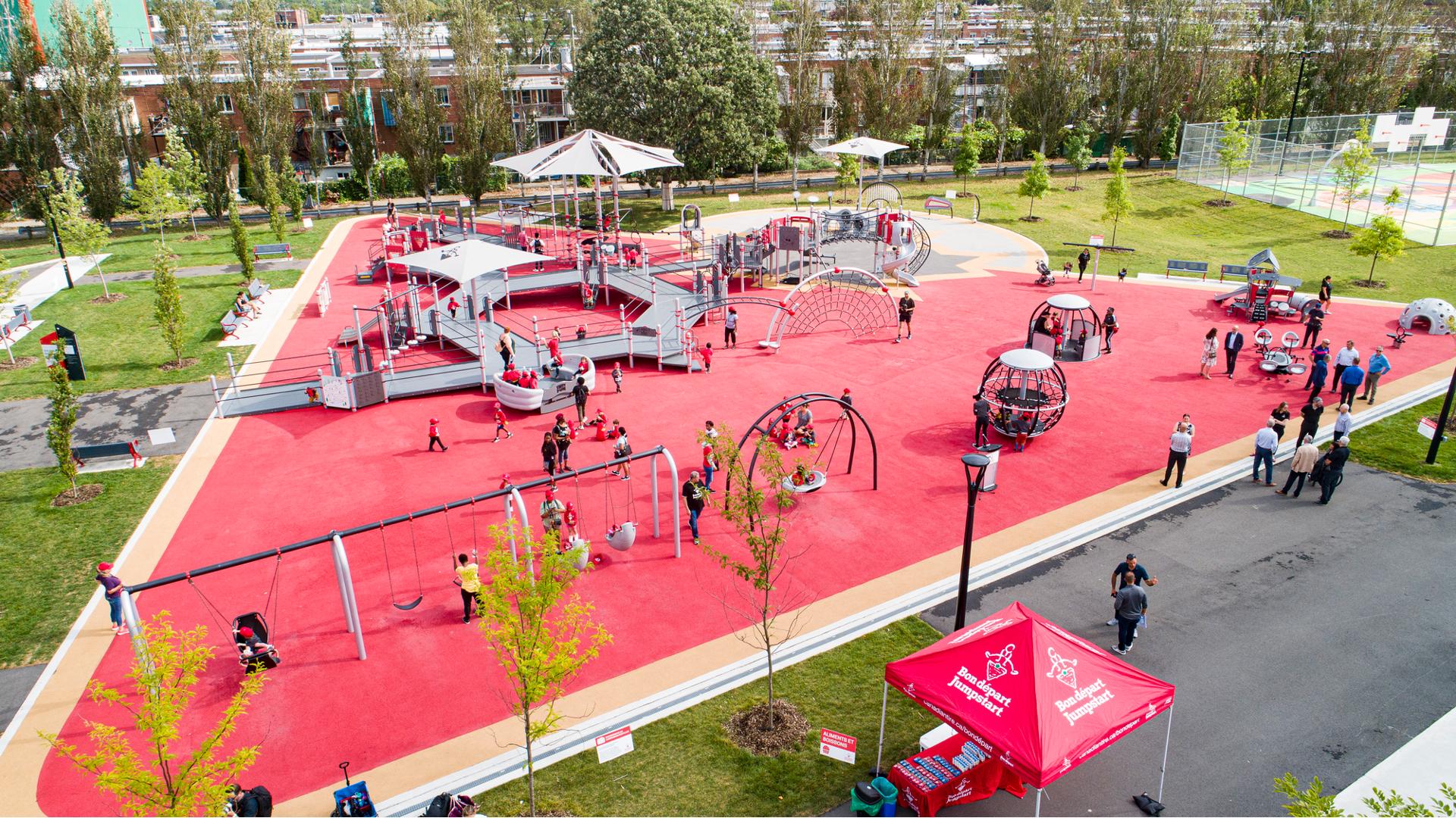 A large triangular shaped inclusive play area contains a play structure made up of accessible ramps. Families play all around the park on swing sets, spinners, and other free standing play activities.