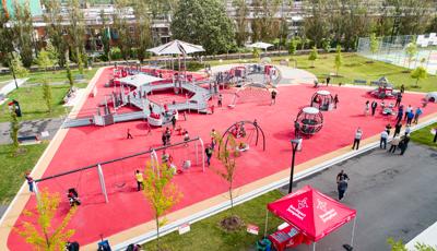 A large triangular shaped inclusive play area contains a play structure made up of accessible ramps. Families play all around the park on swing sets, spinners, and other free standing play activities.