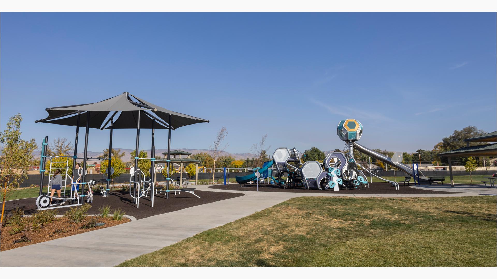 A concrete pathway leads up to and circles around a play area with a play structure made up of hexagonal shaped play pods with connected slides and climbers. Near the play area is a workout area with a large shade with outdoor work out equipment connected to the many shade posts. 