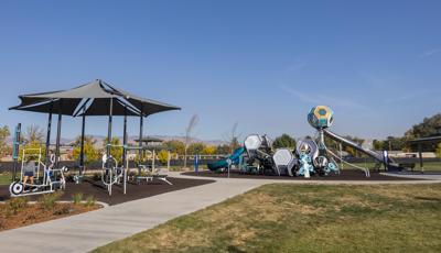 A concrete pathway leads up to and circles around a play area with a play structure made up of hexagonal shaped play pods with connected slides and climbers. Near the play area is a workout area with a large shade with outdoor work out equipment connected to the many shade posts. 