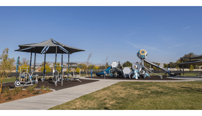 A concrete pathway leads up to and circles around a play area with a play structure made up of hexagonal shaped play pods with connected slides and climbers. Near the play area is a workout area with a large shade with outdoor work out equipment connected to the many shade posts. 