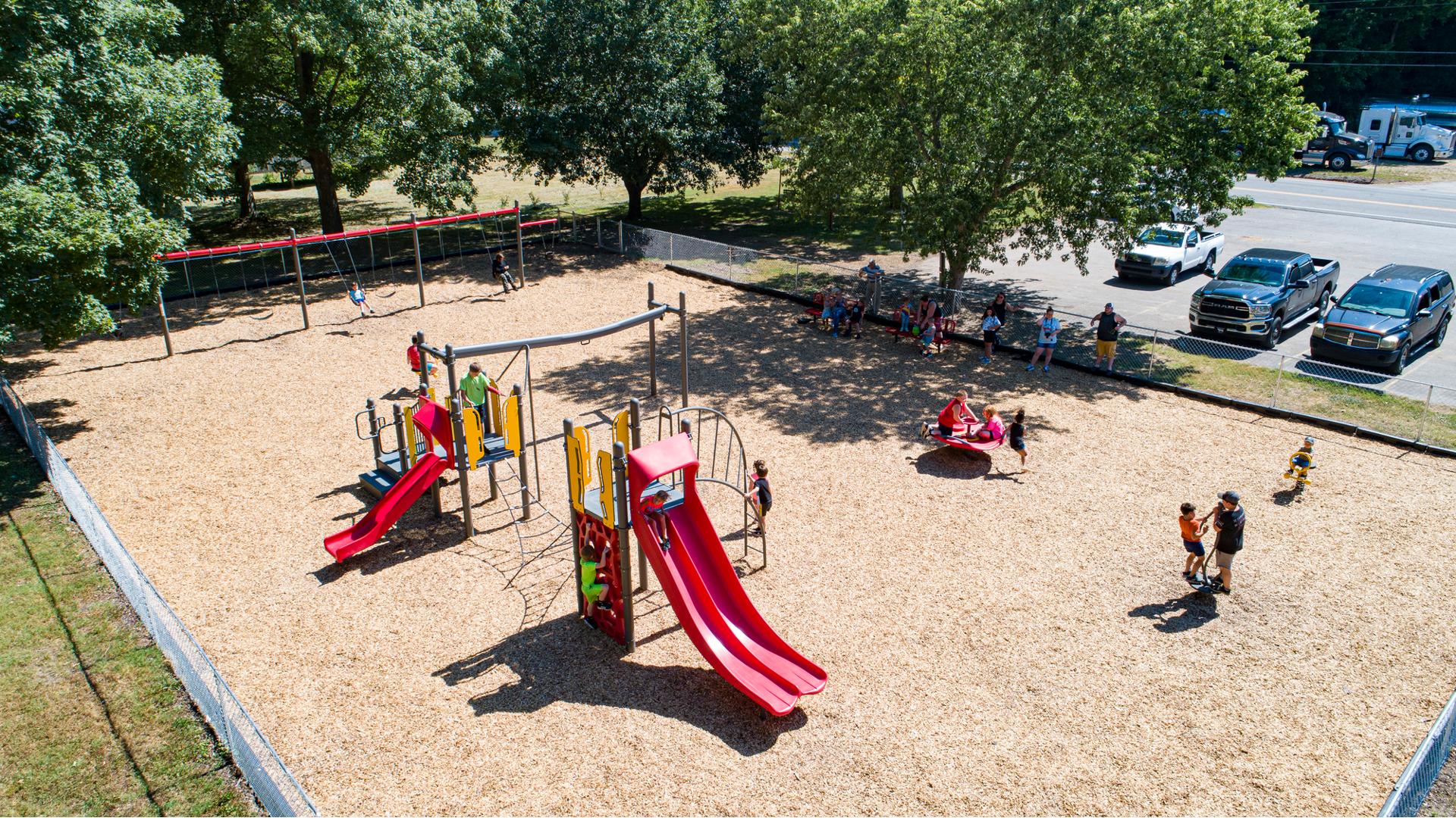 A group of parents sit and stand in the shade of a tree watching as their children play on a play structure filled with slides and climbers. Other children play on spinners and a swing set as well. The large fenced in rectangular play area has a wood chip flooring.