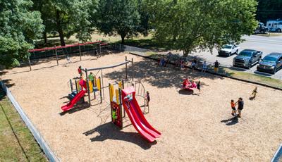 A group of parents sit and stand in the shade of a tree watching as their children play on a play structure filled with slides and climbers. Other children play on spinners and a swing set as well. The large fenced in rectangular play area has a wood chip flooring.