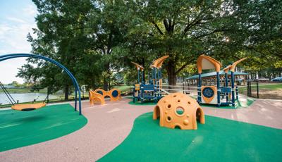 Inclusive PlayBooster playground with custom surfacing and freestanding equipment like the Cozy hut sensory component and Oodle swing with a body of water behind the park and a large tree covering the playground in shade.