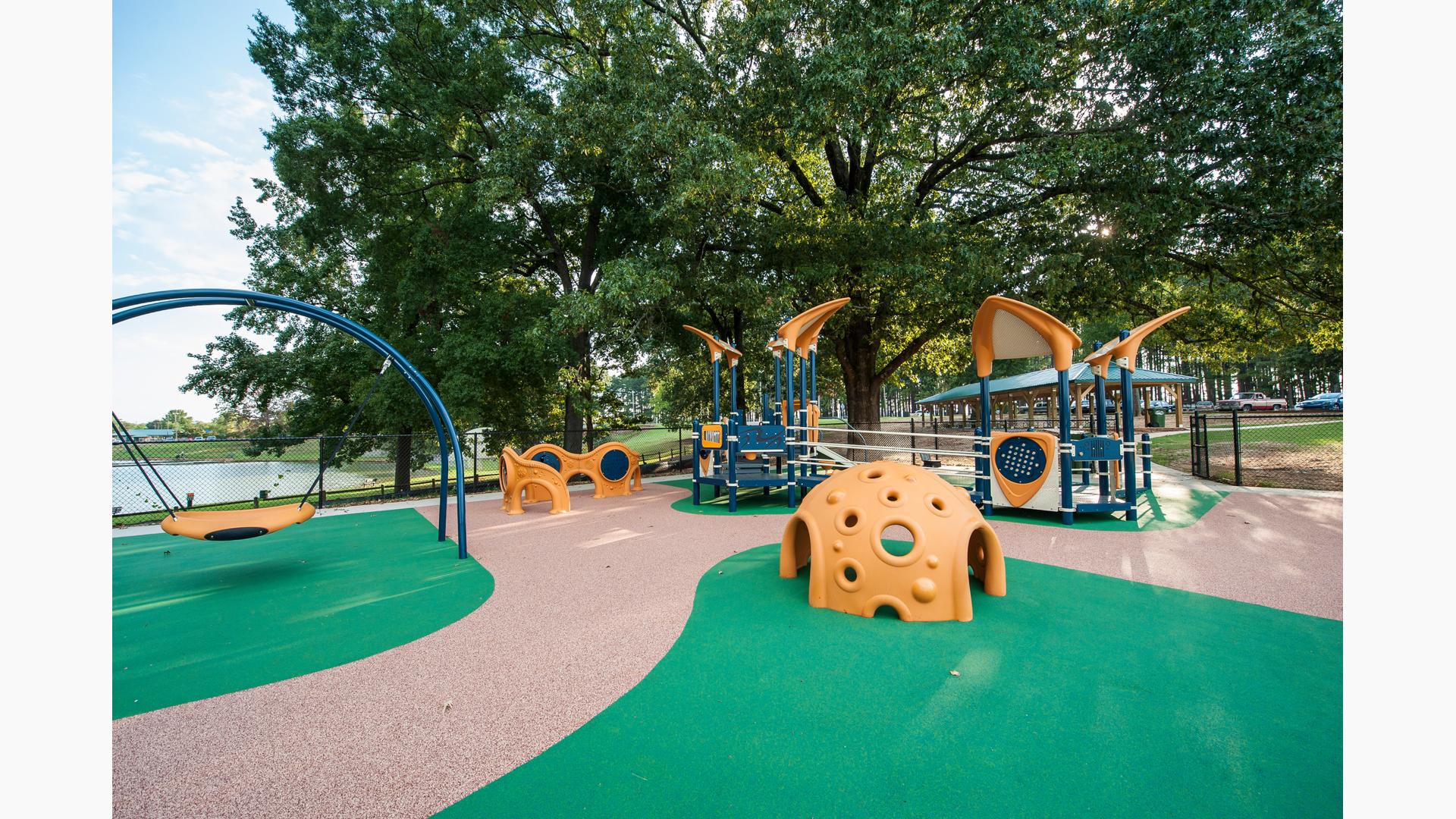 Inclusive PlayBooster playground with custom surfacing and freestanding equipment like the Cozy hut sensory component and Oodle swing with a body of water behind the park and a large tree covering the playground in shade.