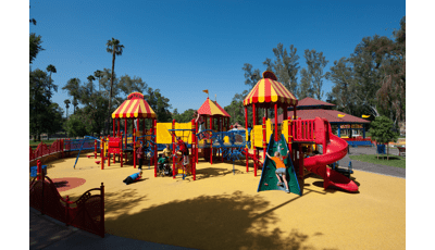 Custom Carnival PlayBooster play structure in Fairmount Carousel Playground give access to children of all abilities. A long ramp reaches around the play structure. A girl in an orange shirt climbs a Conical Climber. A boy lays on a Saddle Spinner. A group of children play on a Chimney Climb.