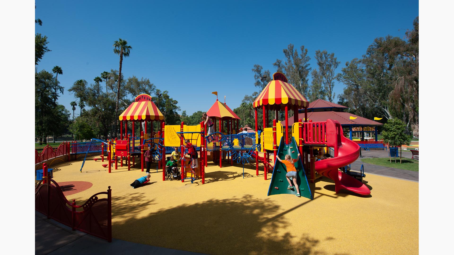 Custom Carnival PlayBooster play structure in Fairmount Carousel Playground give access to children of all abilities. A long ramp reaches around the play structure. A girl in an orange shirt climbs a Conical Climber. A boy lays on a Saddle Spinner. A group of children play on a Chimney Climb.