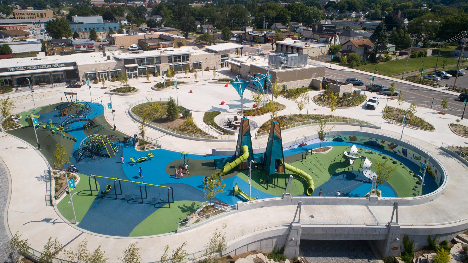 Full elevated view of a large play area with two main modern design tower structures. Other modern curved climbers and play structures are scatter throughout the play area all on safety surfacing colored in different shades of blues and greens.