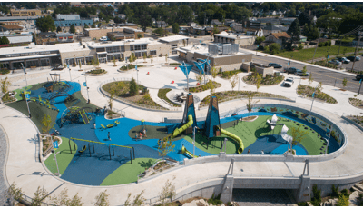 Full elevated view of a large play area with two main modern design tower structures. Other modern curved climbers and play structures are scatter throughout the play area all on safety surfacing colored in different shades of blues and greens.