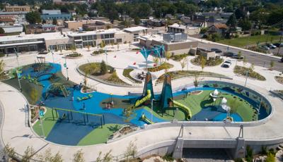 Full elevated view of a large play area with two main modern design tower structures. Other modern curved climbers and play structures are scatter throughout the play area all on safety surfacing colored in different shades of blues and greens.