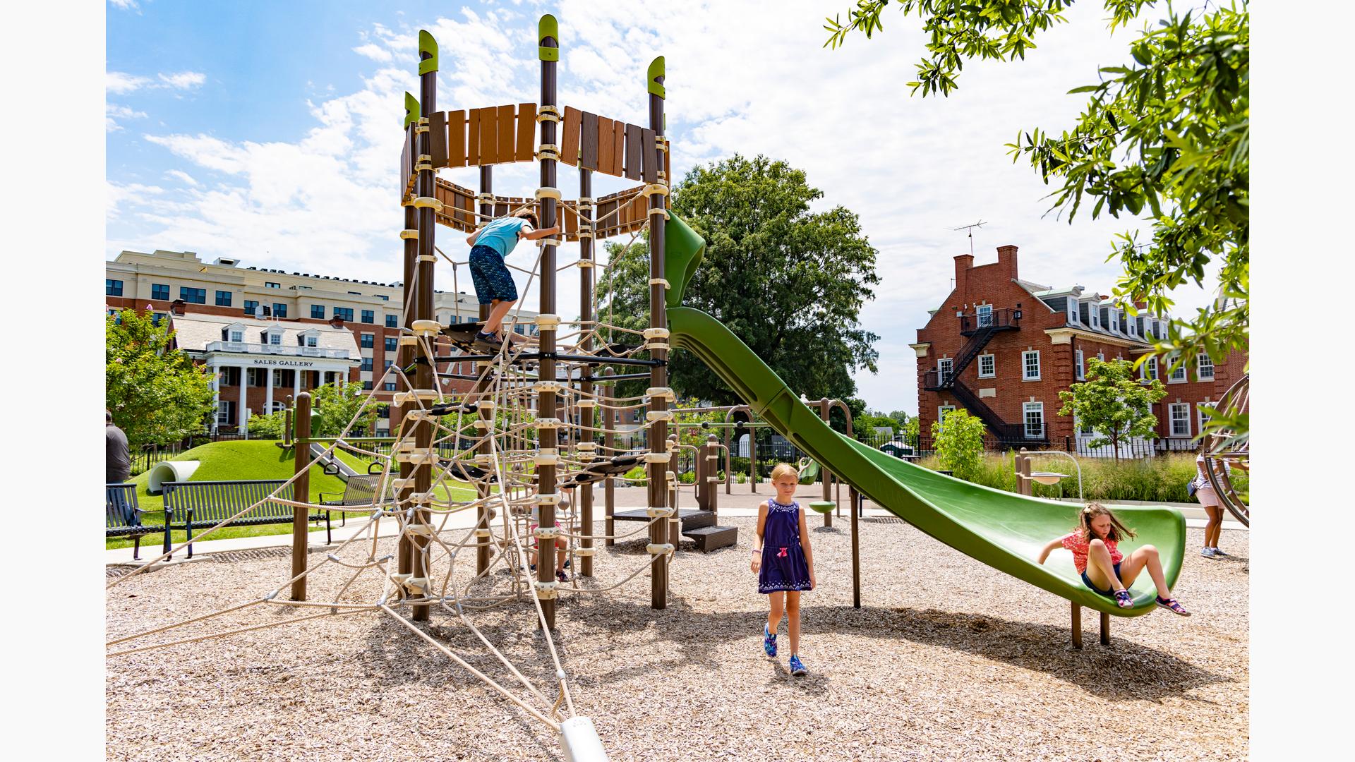 A natural wood-like and net play-based play structure with a girl sliding down a green Alpine Slide.