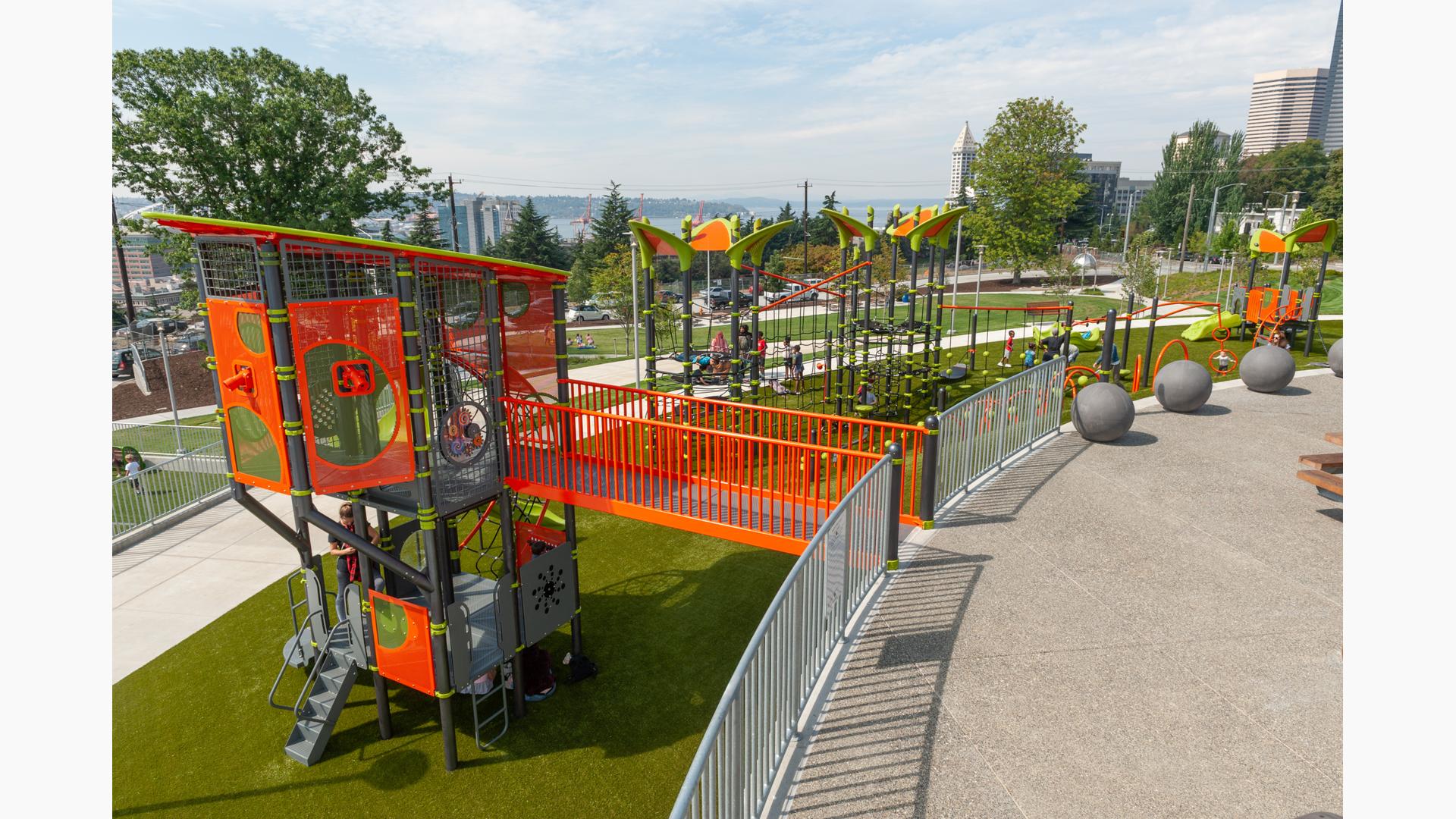 Brightly colored tower structures with ropes and cages to explore. Children playing on playground while parents watch. Green trees and city skyline in the background.