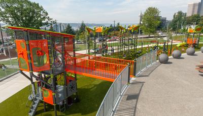 Brightly colored tower structures with ropes and cages to explore. Children playing on playground while parents watch. Green trees and city skyline in the background.