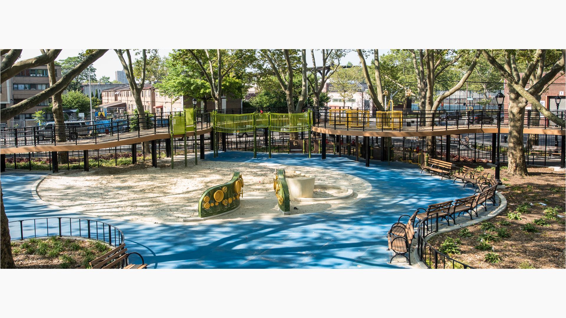 Large trees surround and shade artificale wood planked ramps winding around a large play sand area with custom sensory panel playing walls.