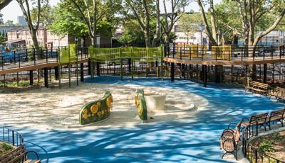 Large trees surround and shade artificale wood planked ramps winding around a large play sand area with custom sensory panel playing walls.
