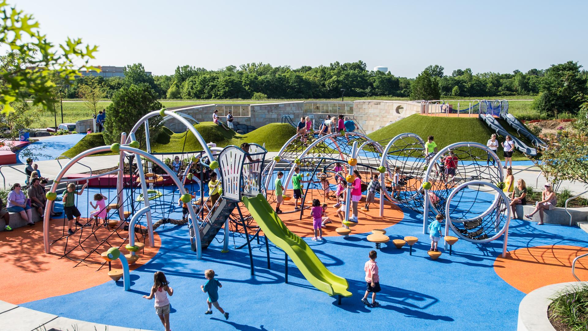A huge custom playscape filled with net climbers for ages 2-12 and a green slide in the foreground and navy blue hill slides in the background. 