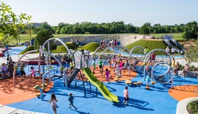 A huge custom playscape filled with net climbers for ages 2-12 and a green slide in the foreground and navy blue hill slides in the background. 