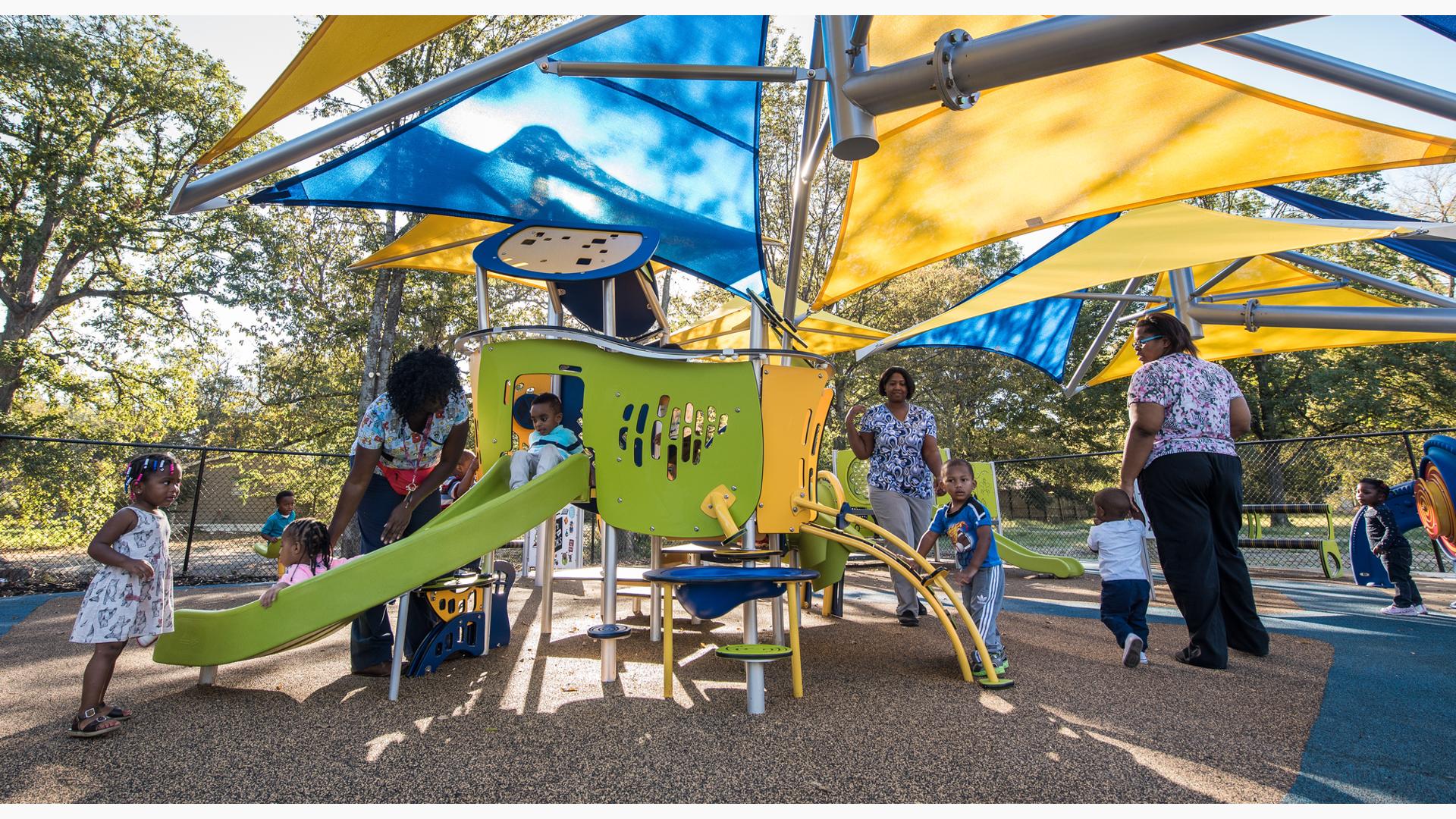 Teachers help these toddlers play on a Smart Play play structure. A girl in a white dress stands at the foot a the Smart Play slide as two children ride down with their teacher's help.