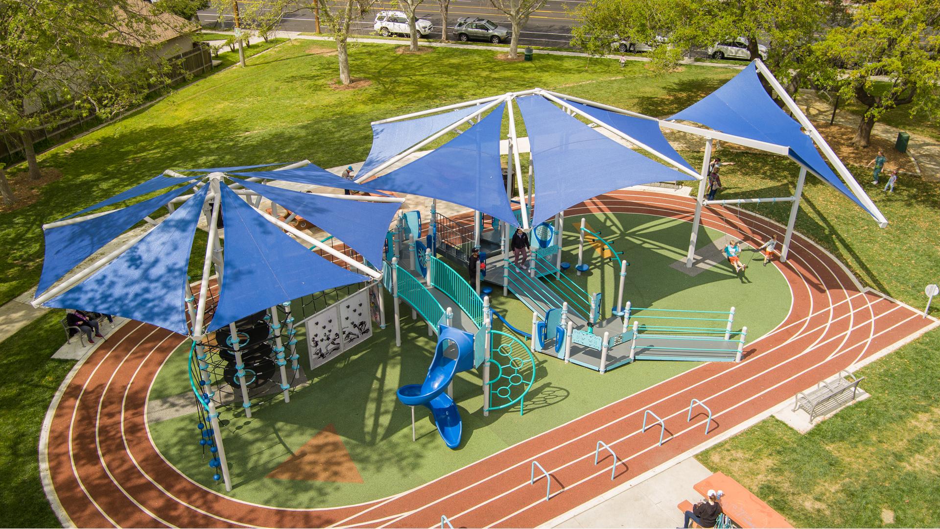 A running track surrounds a large inclusive playground with three large shade systems overhead.