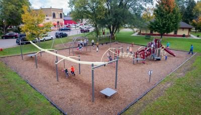 Higher advantage image of a playground in a city park. Kids are running around on the playground and taking turns riding on the zipline and swings. The playground is pale yellow and dark red. 