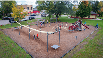 Higher advantage image of a playground in a city park. Kids are running around on the playground and taking turns riding on the zipline and swings. The playground is pale yellow and dark red. 