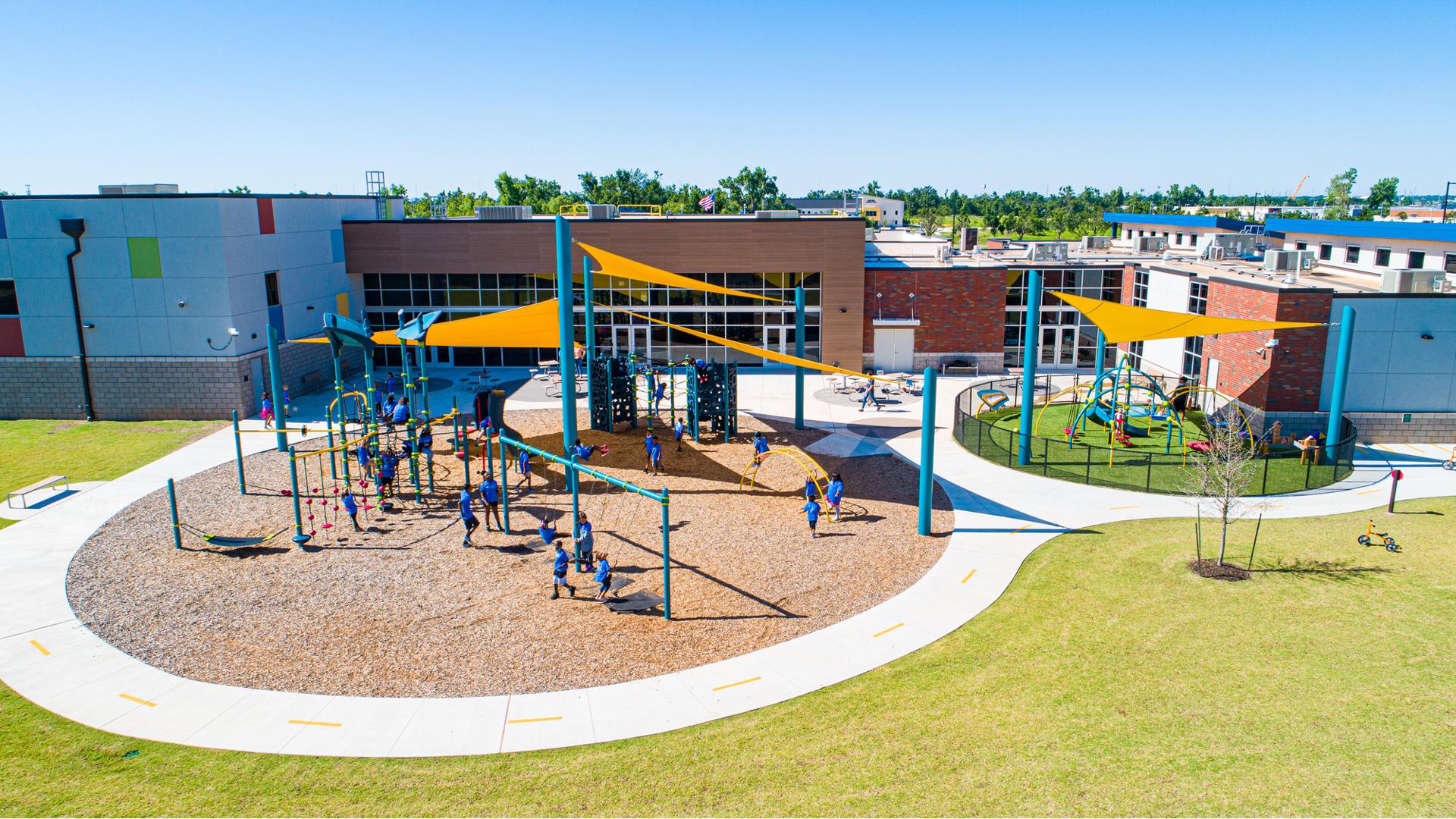 Full elevated view of two separate play areas for all ages of children next to a large school building. The two play areas have large yellow triangular shade sails overhead. 