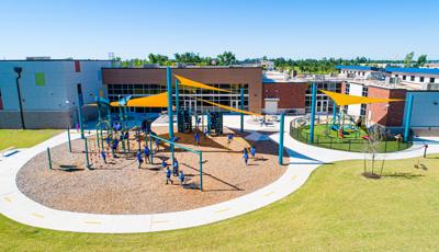 Full elevated view of two separate play areas for all ages of children next to a large school building. The two play areas have large yellow triangular shade sails overhead. 