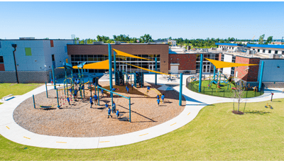Full elevated view of two separate play areas for all ages of children next to a large school building. The two play areas have large yellow triangular shade sails overhead. 