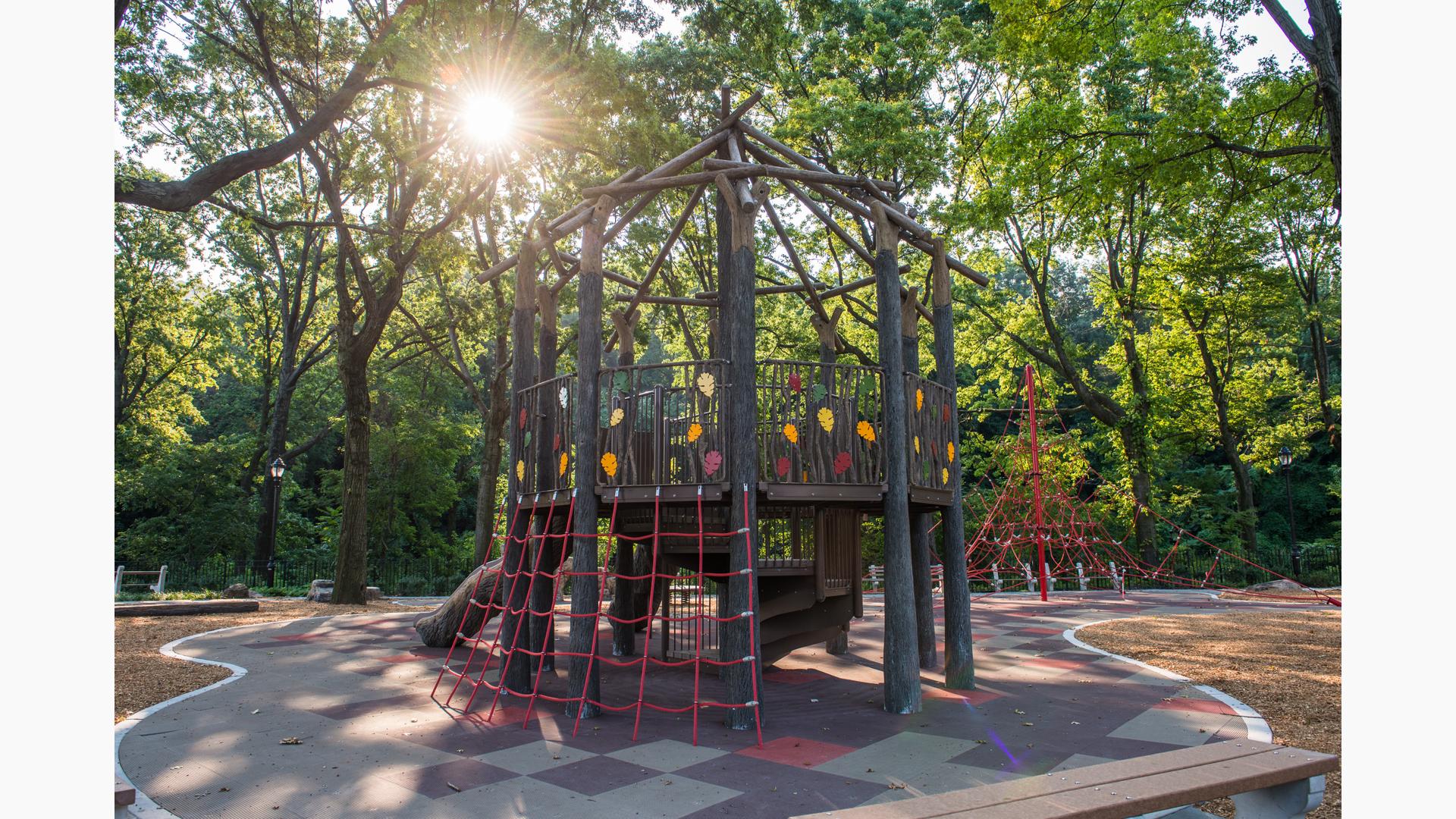 Sun appearing through the trees on a nature-inspired fort-like tower playground . A red triangle-shaped climbing net is in the background. 