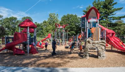 Kids can climb, swing, twirl and slide on a PlayBooster® play structure while an adult in black watches. The Geo Netplex in the background is covered with children at the base.