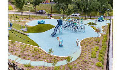 An elevated view of a recreation center park surrounded by a black iron fence. Two separate play A small house like play structure for younger children sits in the background while a larger play area for older children sits in the middle of the park. Multiple large silver arched posts cluster rope and hoop climbers and a slide. The play area slants up a hill with artificial grass, roller slide, and angled climbing rope.