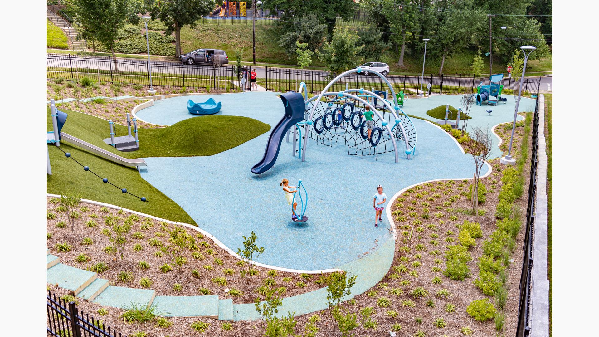An elevated view of a recreation center park surrounded by a black iron fence. Two separate play A small house like play structure for younger children sits in the background while a larger play area for older children sits in the middle of the park. Multiple large silver arched posts cluster rope and hoop climbers and a slide. The play area slants up a hill with artificial grass, roller slide, and angled climbing rope.