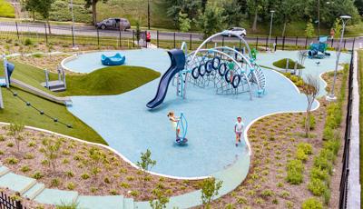 An elevated view of a recreation center park surrounded by a black iron fence. Two separate play A small house like play structure for younger children sits in the background while a larger play area for older children sits in the middle of the park. Multiple large silver arched posts cluster rope and hoop climbers and a slide. The play area slants up a hill with artificial grass, roller slide, and angled climbing rope.