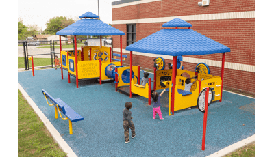 Young children play at two connected play structures made of different play panels set next to a brick building.