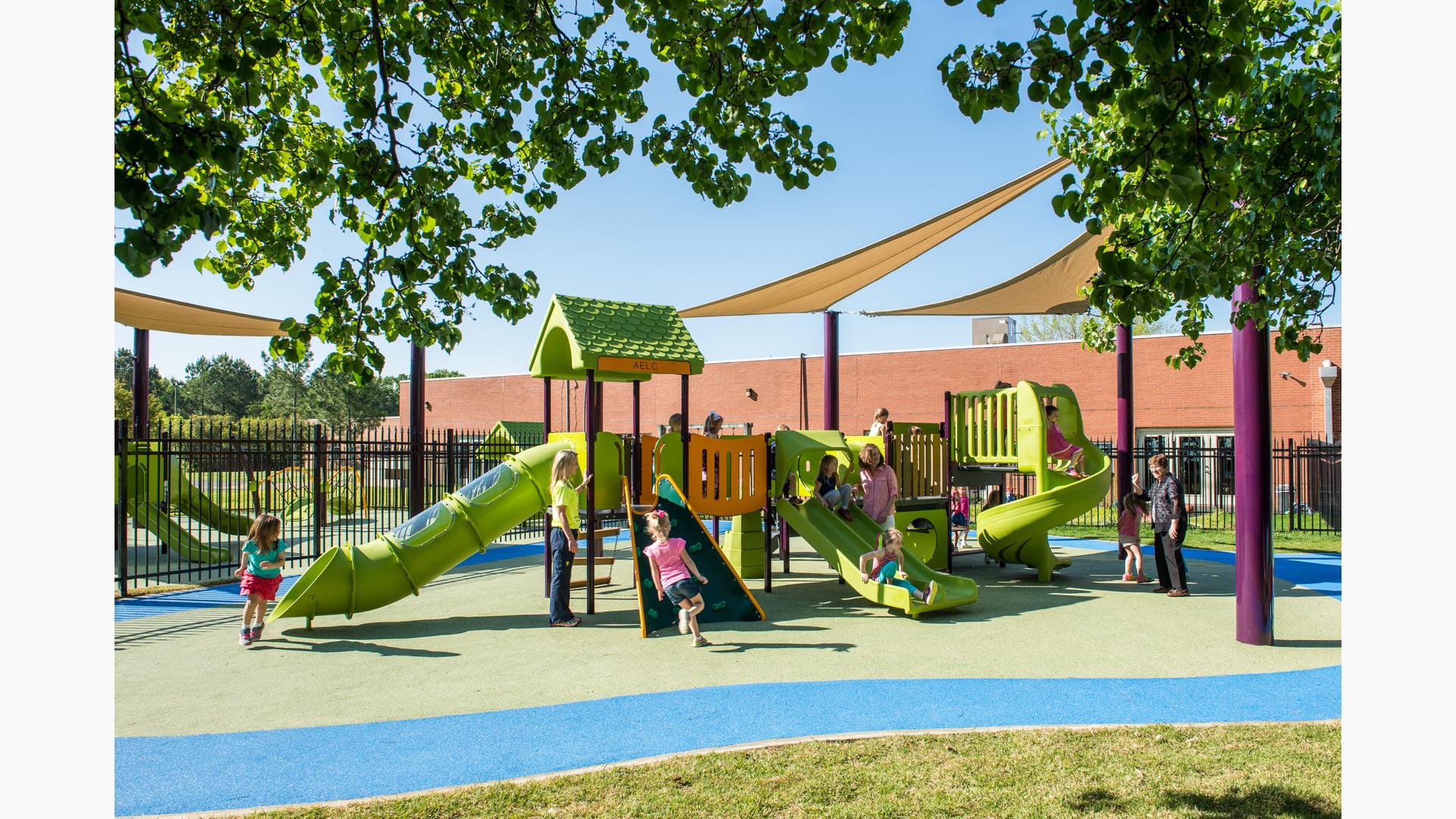 Adults watch as children play on a play structure with green slides, climbers, and bridges surrounded by a black iron fence next to a brick building.
