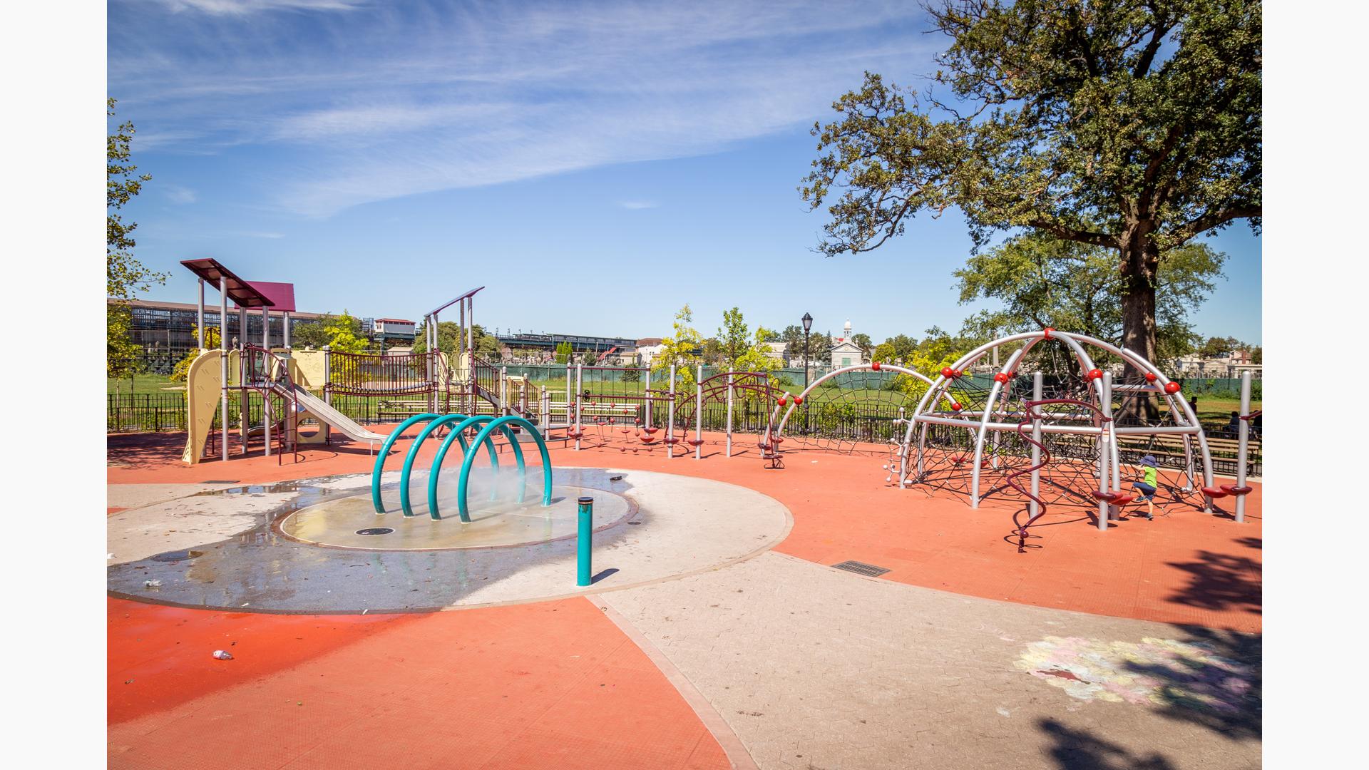 Several steel arched net structures line the edge of the play area and across from a traditional looking slanted flat roof playground. Kids can cool off in the nearby arched splash pad water element located in the center of the play space.