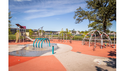 Several steel arched net structures line the edge of the play area and across from a traditional looking slanted flat roof playground. Kids can cool off in the nearby arched splash pad water element located in the center of the play space.