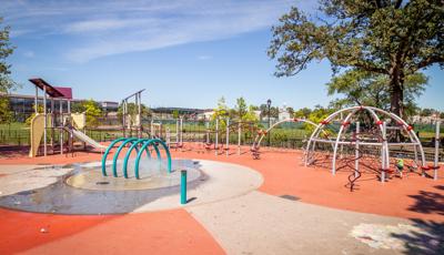 Several steel arched net structures line the edge of the play area and across from a traditional looking slanted flat roof playground. Kids can cool off in the nearby arched splash pad water element located in the center of the play space.