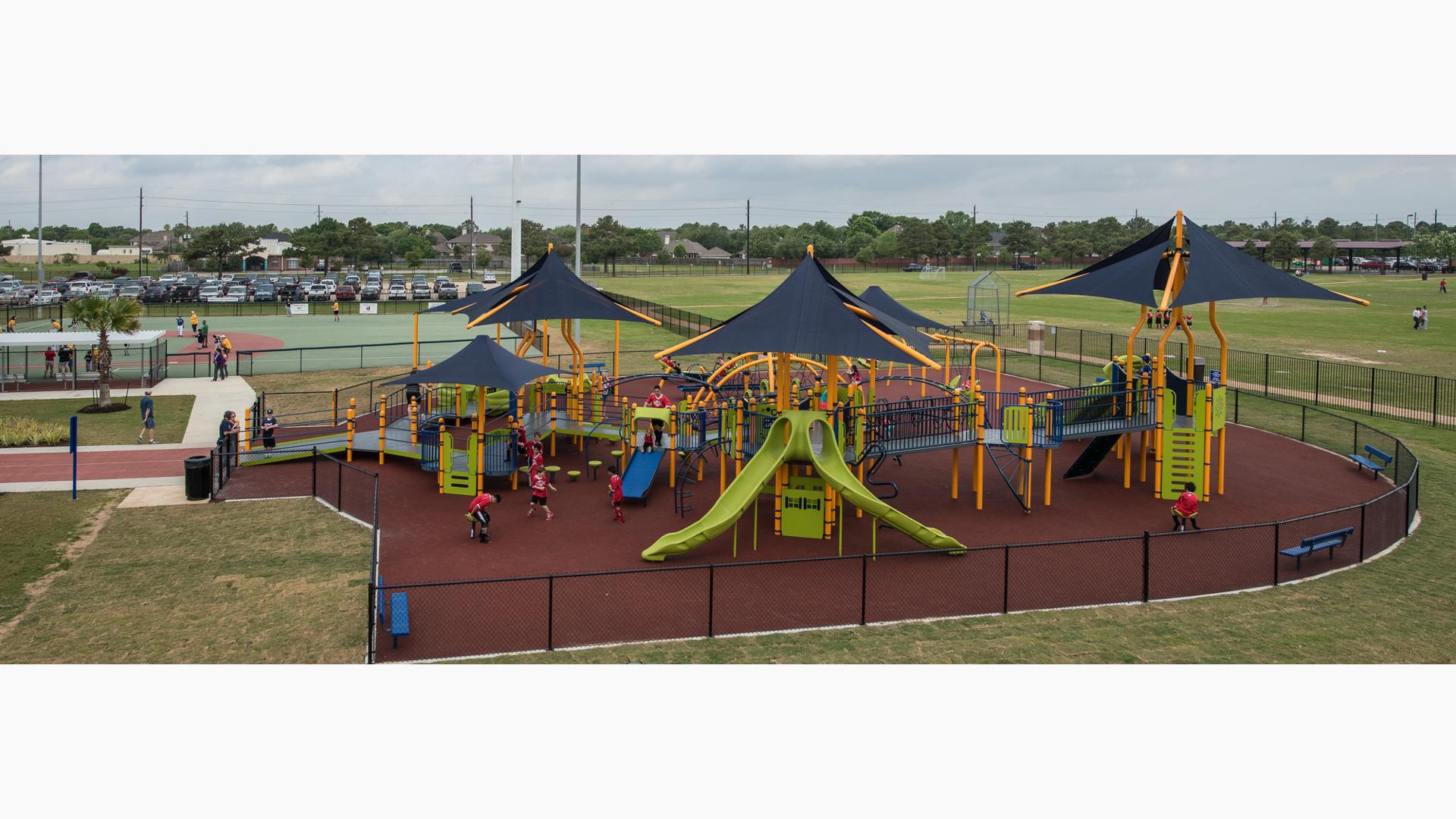 Children of all abilities at play on the Langham Creek Miracle League playground, while baseball training goes on the fields in the background.